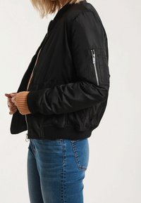 Černá bomber bunda s žebrovanými manžetami a lemem, vybavená kapsou na zip na rukávu a měkkou, hladkou tkaninou.