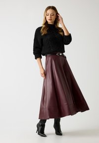 Maglione nero a maglia con motivo a trecce, abbinato a una gonna maxi in ecopelle bordeaux con cintura larga e finitura lucida. Stivaletti neri alla caviglia.