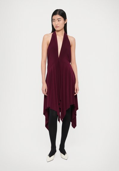 Rochie bordo cu bretele, decolteu adânc în V, tiv asimetric fluid, material ușor, asortată cu ciorapi negri și pantofi albi cu vârf ascuțit.