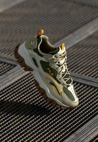 Zapatilla verde y beige con parte superior de malla texturizada, suela de goma, acentos amarillos y logotipo prominente. Cuenta con un cuello acolchado y un sistema de cordones.
