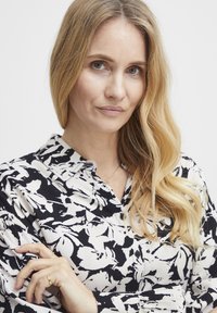 Bijela bluzica s crnim cvjetnim uzorkom, dizajn na kopčanje i meka tekstura. Model ima dugu, valovitu kosu i nosi delikatnu ogrlicu.