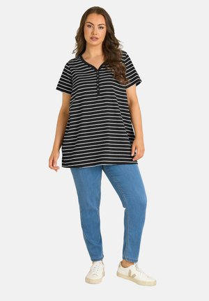 Frau trägt ein schwarz-weiß gestreiftes Kurzarmoberteil, blaue Jeans und weiße Freizeitsneaker, steht vor einem einfarbigen Hintergrund.