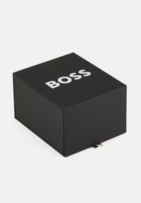 Juodas stačiakampis dėžė su matinio paviršiaus apdaila, viršutiniame paviršiuje pabrėžtu žodžiu "BOSS" baltomis paryškintomis raidėmis. Minkštas paviršius su auksiniais akcentais.