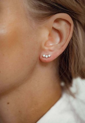 Drei kleine weiße Perlenohrringe, die in einer silbernen Metallbasis eingefasst sind und in einem linearen Muster angeordnet sind. Zu sehen sind sie in einer Nahaufnahme eines Ohrläppchens.