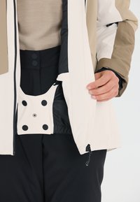 Giacca impermeabile con un esterno crema e beige, caratterizzata da dettagli neri. L'interno include una tasca sicura con bottoni a pressione.