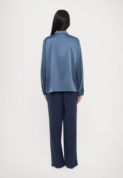 Mujer con cabello largo y oscuro, vistiendo una blusa de satén azul de corte holgado y pantalones anchos de color azul marino, de pie de espaldas sobre un fondo blanco.