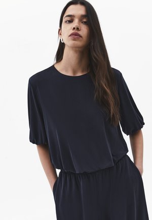 Vrouw met lang donker haar die een loszittende zwarte blouse met geplooide mouwen en bijpassende zwarte broek draagt, staand met handen in de zakken.