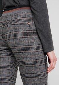 Gros plan sur une personne portant un pantalon à carreaux gris avec une poche arrière et un petit logo rond en métal, associé à un haut à manches longues gris foncé.