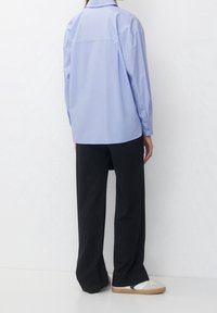 Persona che indossa una camicia oversize azzurro chiaro, pantaloni neri a righe verticali larghi e sneakers bianche con suole in gomma, in piedi contro uno sfondo bianco.