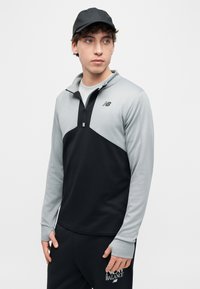 Νέος άνδρας που φοράει μαύρο και γκρι μπουφάν New Balance με φερμουάρ μέχρι τη μέση, μαύρη φόρμα New Balance και μαύρο καπέλο, κοιτάζοντας προς τα δεξιά.