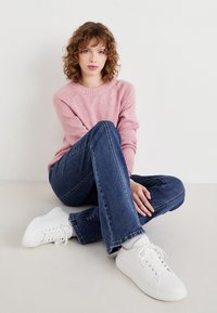 Young woman with curly hair sitting on floor, wearing pink sweater, blue jeans, white socks, and white sneakers against plain white background.