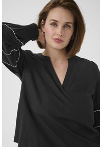 Schwarze Bluse mit V-Ausschnitt und puffärmeligen Ärmeln, die mit gerafften Akzenten versehen sind. Glatte Textur mit weißen Details entlang der Ärmel.