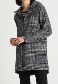 Mujer vistiendo un abrigo con capucha de cuadro gris y negro, con mangas largas, combinado con pantalones negros.