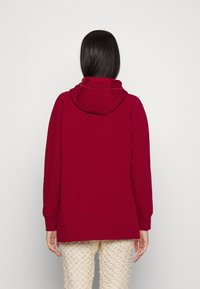 Sudadera burdeos con una gran capucha, corte relajado y puños acanalados. Tejido suave, diseño minimalista sin logotipos ni patrones visibles.
