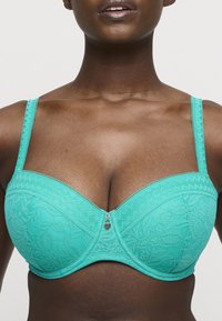 Turkos spets-bh med byglar, blommönster, justerbara axelband och en dekorativ hjärtcharm i mitten. Slät textur.