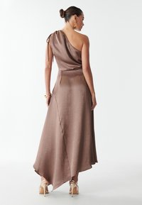 Abito in satin marrone con design a spalla singola, caratterizzato da un dettaglio arricciato e un'elegante orlo asimmetrico. Indossato con tacchi a strappo.