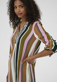 Robe rayée présentant des lignes verticales en vert, rose, noir et blanc ; dotée d'un col en V et de manches retroussées avec des accents de boutons.