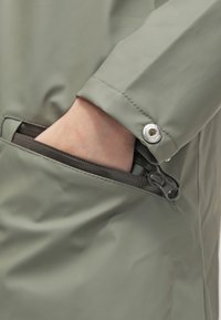 Světl zelená voděodolná bunda se zipovou kapsou. Detailní záběr ruky v kapse; obsahuje stříbrný patent a hladkou texturu.