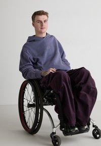 Hanorac purpuriu cu un logo în spirală, pantaloni largi întunecați, așezat într-un scaun cu rotile cu un cadru argintiu și roți cu accente roșii. Fundal neutru.