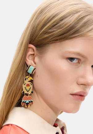 Zbliżenie kobiety z prostymi, blond włosami, noszącej kolorową, koralikową kolczyk w kształcie papugi na lewym uchu.