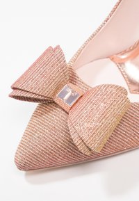 Chaussure à talon haut rose texturée avec un bout pointu, ornée d'un grand nœud et d'un accent métallique à l'avant. Tissu élégant et scintillant.