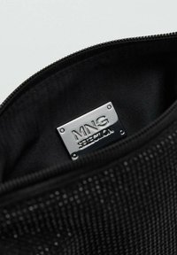 Interno di una borsa nera con cerniera che presenta un'etichetta rettangolare argentata impreziosita con la dicitura "MNG SELECTION." Tessuto liscio con texture e lucentezza sottile.