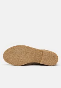Semelle de chaussure beige clair en matériau texturé semblable à du caoutchouc. Forme ronde avec un léger talon et un détail de marque. Design plat adapté à un usage décontracté.