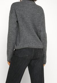 Grauer gestrickter Pullover mit gerippter Struktur, hohem Kragen, lockerer Passform und herunterfallenden Schultern, kombiniert mit hochgeschnittenen dunklen Jeans.