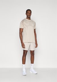 Ανοιχτό μπεζ μπλουζάκι με υφή, σε συνδυασμό με ανοιχτόχρωμες σορτς. Μοντέλο που φοράει λευκά αθλητικά παπούτσια και κάλτσες μέχρι τον αστράγαλο, σε φόντο χωρίς σχέδιο.