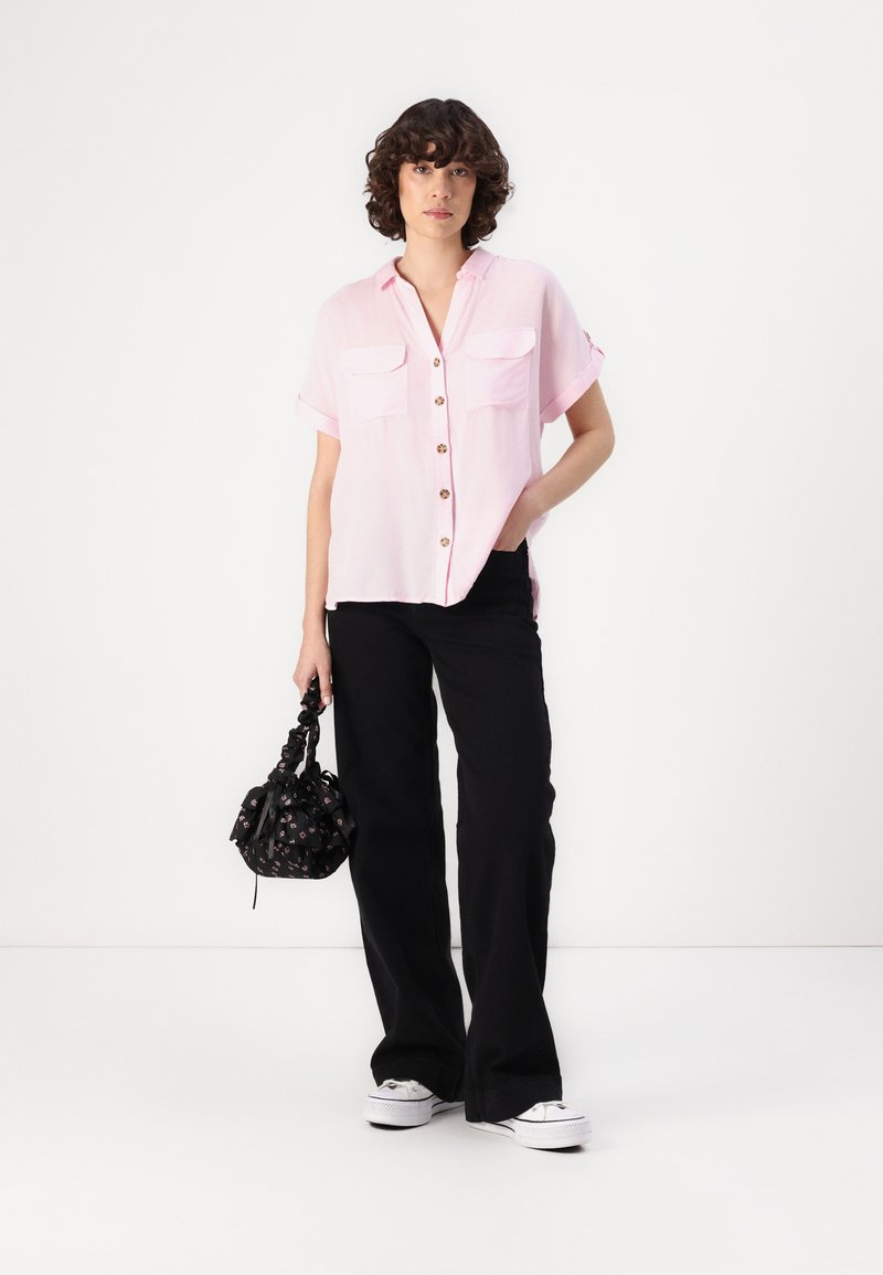 Chemise rose pâle à manches courtes avec deux poches avant et des boutons marron, associée à un pantalon noir à jambes larges et un sac à main noir à motifs.