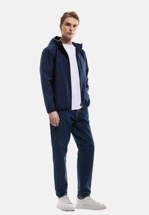 Marineblå vandafvisende jakke med hætte, frontlynlås og elastiske manchetter, båret over en hvid skjorte og parret med blå jeans og hvide sneakers.