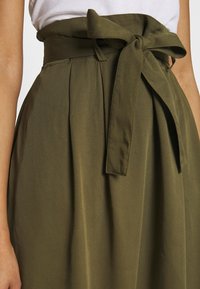 Olivenfarbene Rock mit hoher Taille und einer gebundenen Schleifenoptik. Der Stoff hat eine glatte Textur und weist Falten auf, die zusätzlich Volumen und Form verleihen.