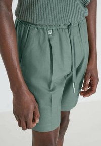 Pantaloni corti verdi, leggeri, con vita elasticizzata, chiusura con cordino e tasche laterali; davanti plissettato e texture liscia.