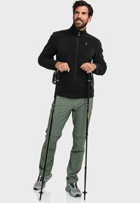 Chaqueta de forro polar negra con cuello alto, pantalones de senderismo verdes y zapatos de senderismo grises. Sosteniendo bastones de trekking negros con detalles amarillos.