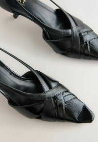 Black heeled slingback shoes with a pointed toe, made of textured patent leather, featuring crisscross straps and a sleek silhouette.