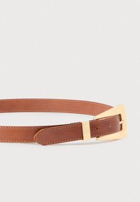 Καφέ δερμάτινη ζώνη με χρυσή τρίγωνη αγκράφα και αντίστοιχο χρυσό θηλάκι σε απλό ανοιχτό γκρι φόντο.