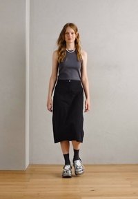 Haut sans manches gris, jupe midi noire avec boutons sur le devant, chaussettes noires, sabots en argent, debout sur un sol en bois contre un mur neutre.
