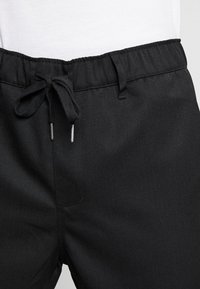 Černé tepláky s elastickým pasem a šňůrkou, mají hladkou texturu a dvě přední kapsy. Jednoduchý, pohodlný design.