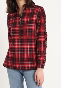 Rood en zwart geruit zip-up top met een staande kraag en decoratieve zijruche langs de mouwen; gemaakt van zachte, geruite stof.