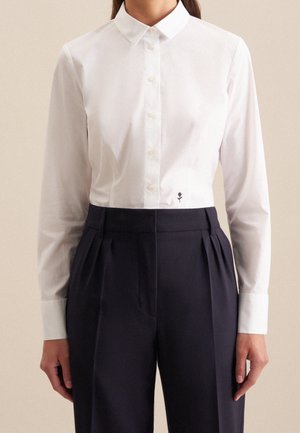 Personne portant une chemise blanche ajustée à boutons avec une petite fleur noire brodée près de la taille, associée à un pantalon noir taille haute.