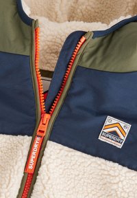 Fermeture éclair partiellement ouverte sur une veste texturée en laine avec des panneaux en tissu bleu marine et vert olive, une fermeture éclair orange vif et un patch logo Superdry Mountain Sport.
