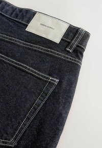 Tumed sinine teksariie, kontrastõmblustega ja halli sildiga, millel on kiri "DENIM SUPPLY" vöökoht, kahte tagataskut.