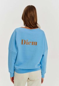 Jasnoniebieska bluza w luźnym kroju, z dużym brązowym nadrukiem "Diem" na plecach oraz ściągaczami przy mankietach i dolnym brzegu. Miękka mieszanka bawełny.