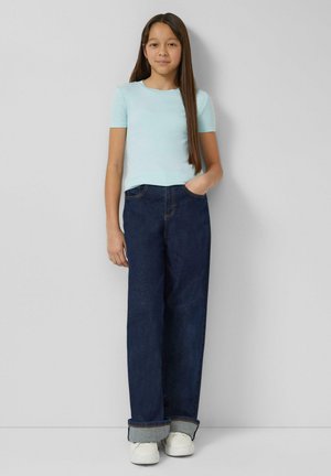 Meisje met lang, stijl haar draagt een lichtblauw T-shirt met korte mouwen, donkerblauwe wijd uitlopende spijkerbroek met omslagen en witte sneakers, staand tegen een grijze muur.