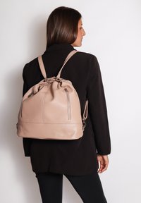 Zaino in pelle rosa con apertura superiore a coulisse, due cerniere laterali e spallacci regolabili, indossato sulla schiena. Texture liscia con hardware minimale.