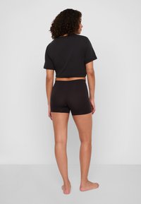 Svart kortärmad t-shirt i kombination med åtsittande svarta shorts. Modell vänd bort, vilket visar det släta tyget och den enkla designen. Enfärgad bakgrund.