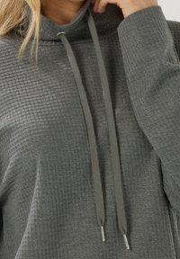 Harmaa huppari, jossa on teksturoidusta, vohvelimaisesta kankaasta valmistettu pinta. Varustettu korkealla kauluksella, nyöreillä ja hopeansävyisillä yksityiskohdilla.