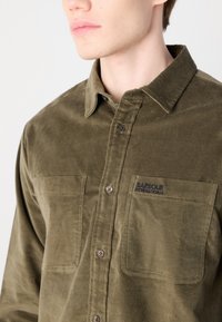 Olive green, button-up shirt made of soft fabric. Features two chest pockets, a collar, and embroidered logo on the left pocket.