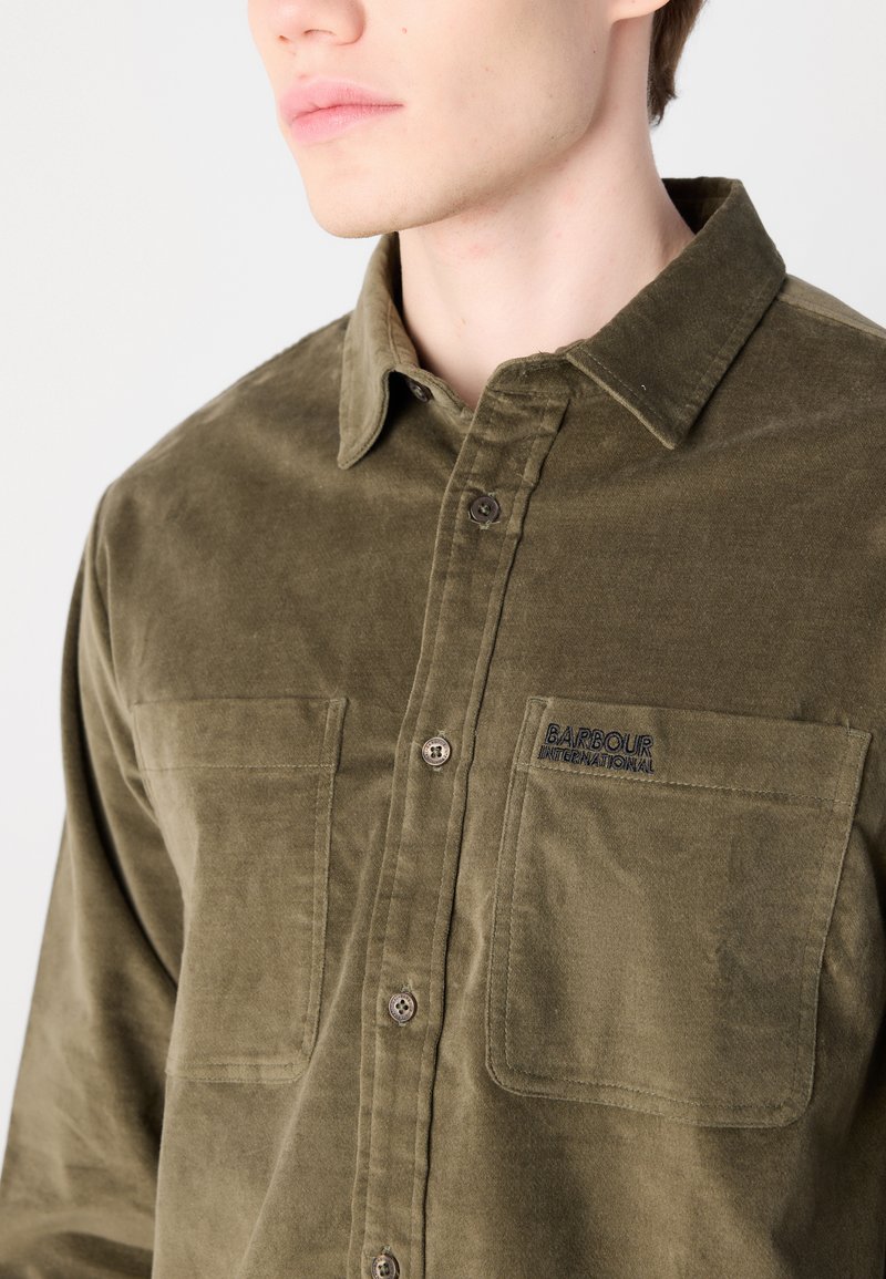 Olijfgroen, knoopshirt gemaakt van zachte stof. Bevat twee borstzakken, een kraag en een geborduurd logo op de linkerklep van de zak.
