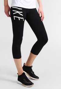 Leggings capri neri con logo "NIKE" bianco sul lato sinistro, abbinati a scarpe da ginnastica nere con dettagli beige.